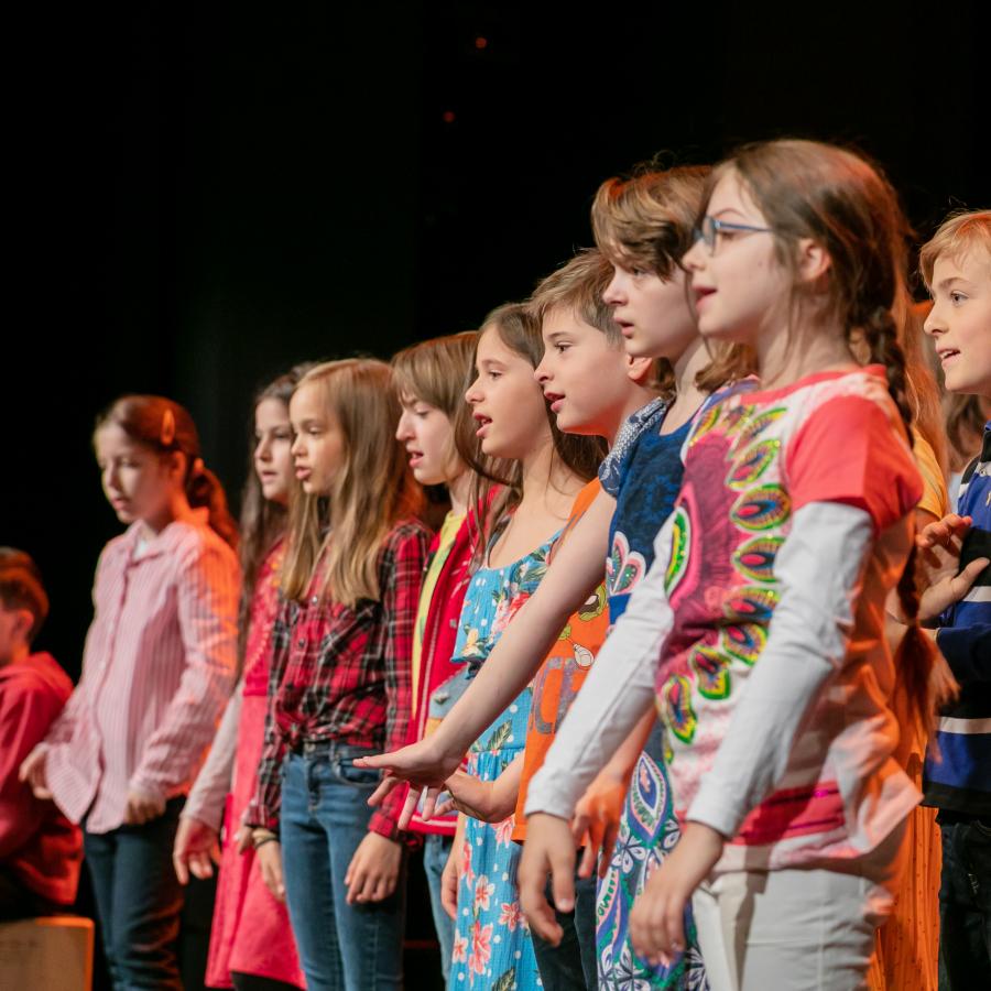 Auf einer Bühne steht eine Schulklasse. Alle Kinder stehen in Reihen und singen ein Lied, das sie zusammen eingeübt haben.  