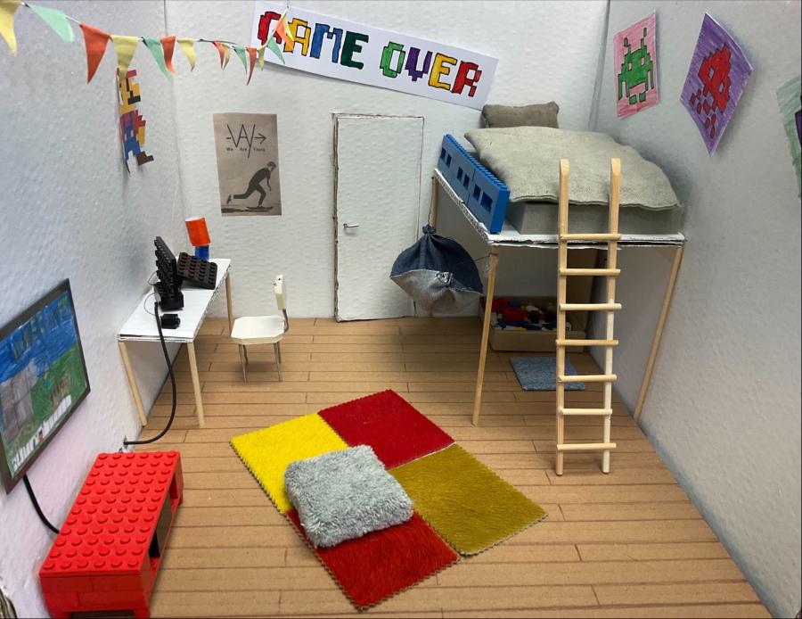 Zu sehen ist ein Modell aus dem Kunstunterricht. Die Kinder sollten ein Modell ihres Kinderzimmers nachbilden. Dieses Zimmer hat ein Hochbett, einen bunten Teppich, Poster und einen Fernseher. 